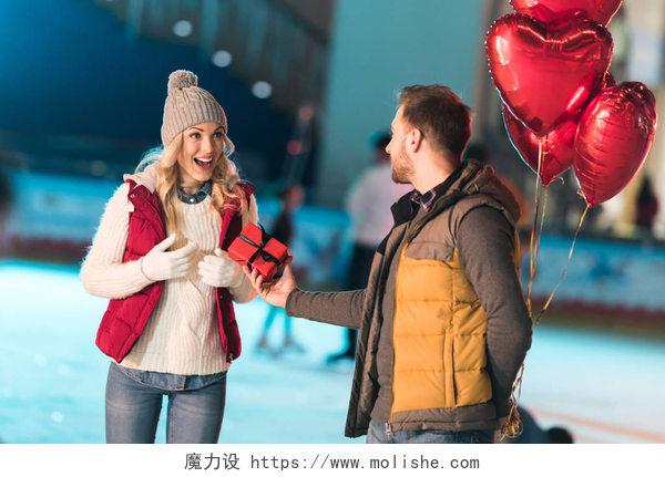 年轻人抱着心形气球和赠送礼物盒给兴奋的女朋友在溜冰场情人节